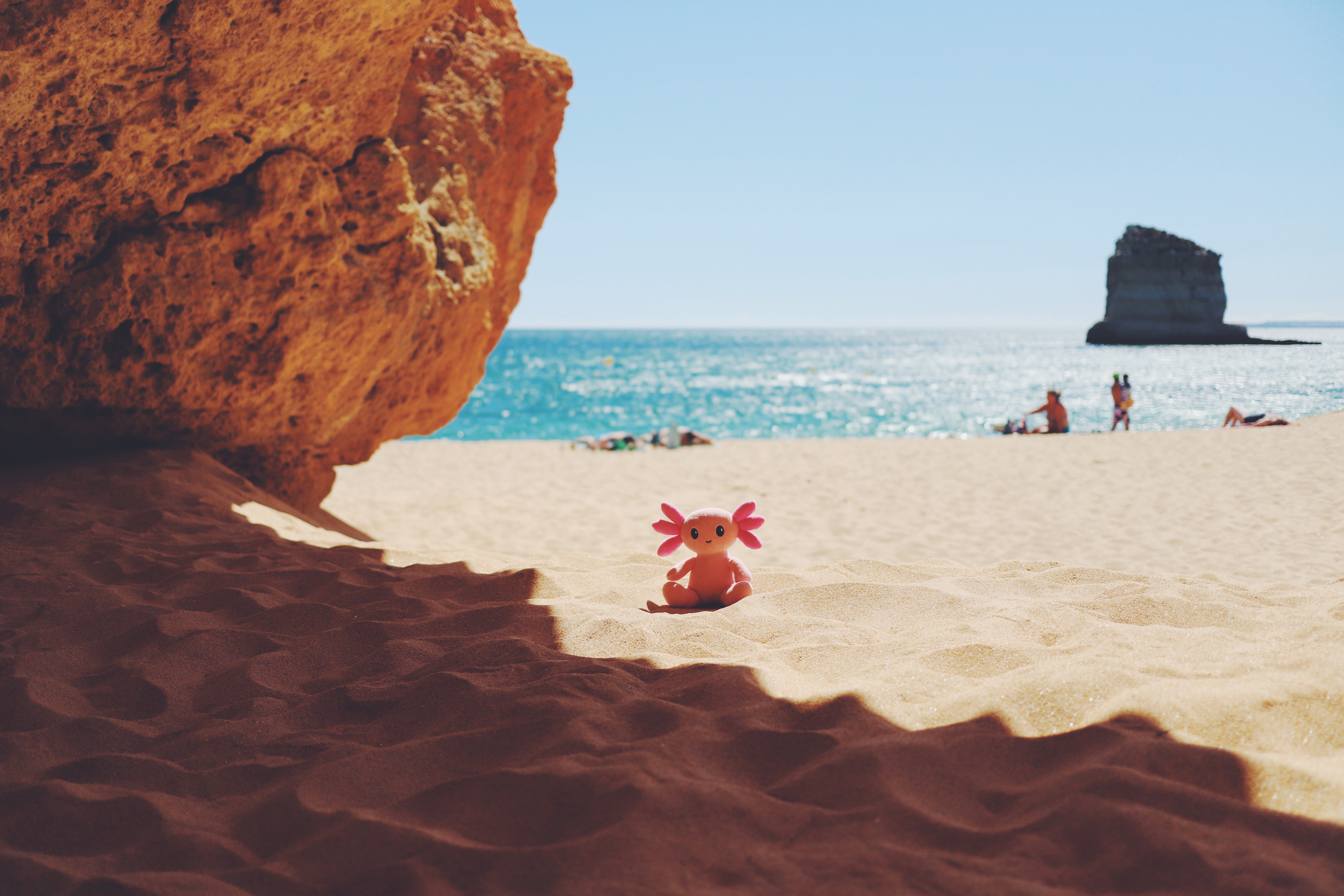 A small pink and orange axolotl plush toy with pink gils sits on a sandy beach under a large rock, with the blue ocean, sky, and a sea stack visible in the background.
